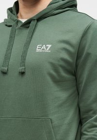 Mikina s kapucňou v tmavozelenej bavlnenej zmesi, s predným vreckom, textúrovanými šnúrkami a malým bielym logom na hrudi.