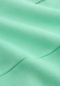 Close-up van mintgroene sweatshirtstof met een gladde textuur, subtiele rekbaarheid en een lichtgeprinte, zichtbare logo in het bovenste gedeelte.