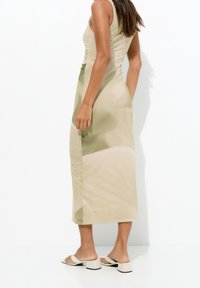 Robe longue et ajustée de teinte beige clair, avec des côtés froncés et une texture lisse et soyeuse. Portée avec des sandales simples et plates.