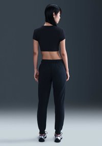 Kvinne står med ryggen til iført en svart cropped t-skjorte, svarte joggebukser og sølvfargede sneakers mot en mørkegrå bakgrunn.