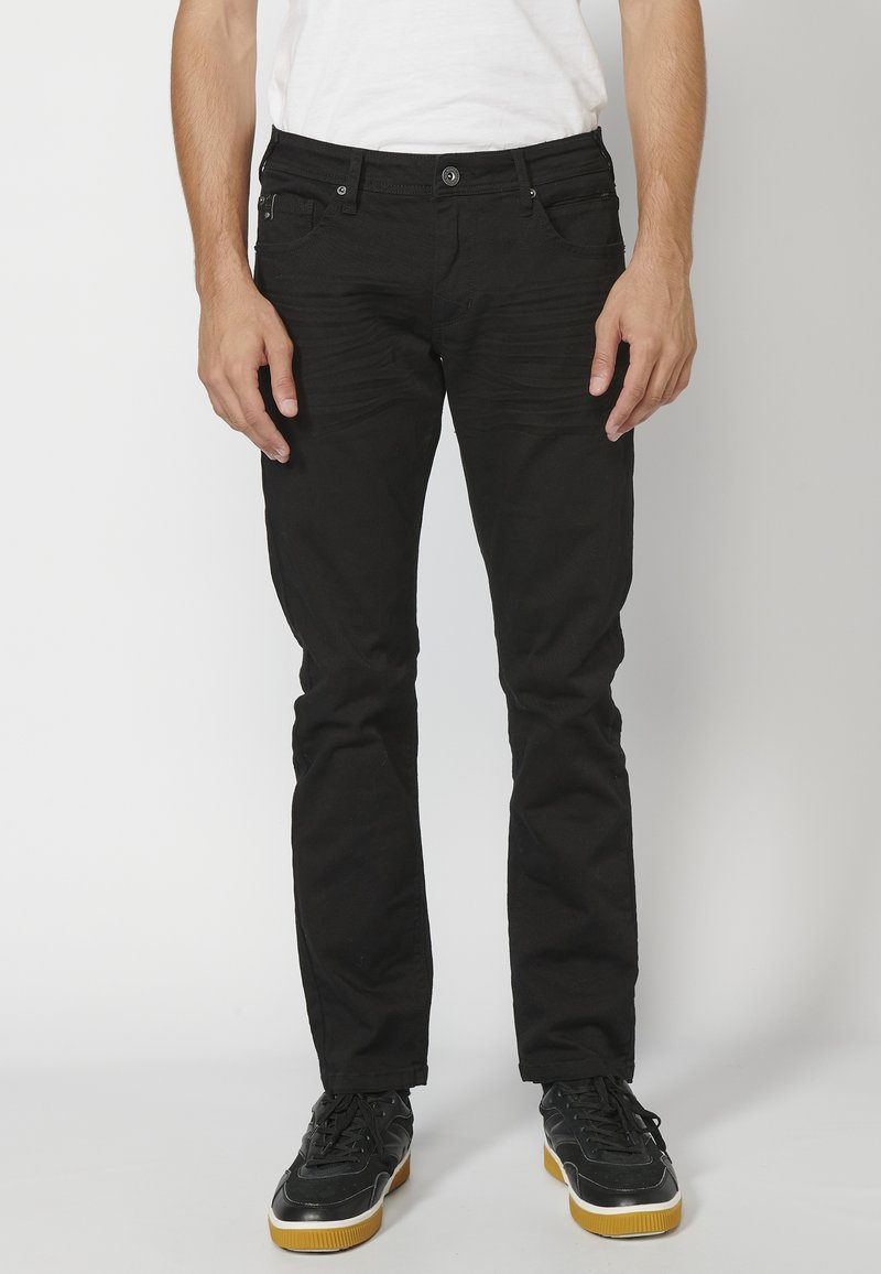 Fekete, slim fit farmernadrág texturált felülettel, öt zsebbekkel és gombos záródással. Fekete sportcipővel párosítva, amelynek gumi talpa van.