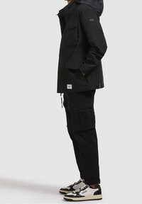 Personne portant un pantalon cargo noir, une veste à capuche noire avec des poches et des baskets noir et blanc, debout de profil les mains dans les poches.