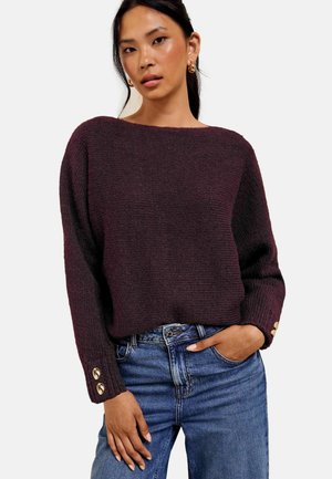 Frau trägt einen dunkelvioletten gestrickten Pullover mit goldenen Knöpfen an den Ärmeln und blaue Jeans, steht mit einer Hand in der Tasche und neutralem Gesichtsausdruck.