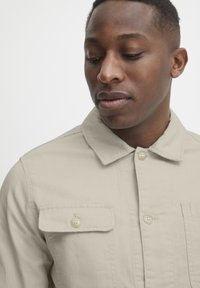 Beige button-up jacket with a collar, featuring two chest pockets and a smooth texture. Visible buttons are off-white.