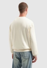 Man met kort haar, gekleed in een effen crèmekleurig sweatshirt en lichtblauwe jeans, staande met zijn rug naar de camera tegen een grijze achtergrond.
