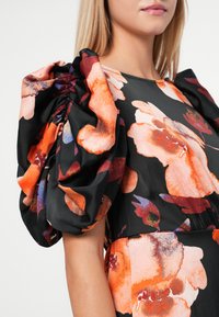 Μαύρο φόρεμα με μεγάλα φουσκωτά μανίκια, διακοσμημένο με λουλούδια σε αποχρώσεις πορτοκαλιού και μπλε. Λείος υφασμάτινος χαρακτήρας με πτυχώσεις στα μανίκια.