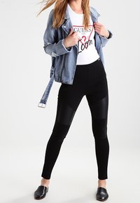 Jeansjacka med slitna detaljer, vit grafisk t-shirt, svarta leggings med paneldetaljer och svarta slip-on skor.