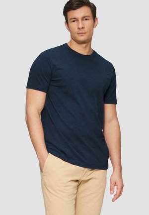 Mann in marineblauem kurzärmeligem T-Shirt und beigen Hosen, der mit einer Hand in der Tasche vor einem einfarbigen hellgrauen Hintergrund steht.