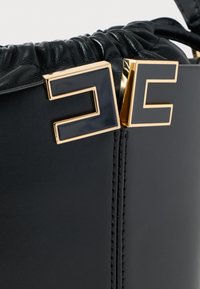 Μαύρη δερμάτινη τσάντα χειρός με πτυχωτή υφή, που διαθέτει χρυσή γεωμετρική λεπτομέρεια κλεισίματος. Λείο επιφάνεια με ραμμένα διακοσμητικά στοιχεία.