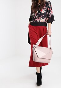 Borsa geometrica rosa con un design a contrasto di rosa chiaro, indossata a tracolla. Modello stilizzato con una blusa nera a fiori e pantaloni rossi a gamba larga.