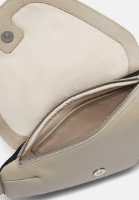 Jasnobeżowa torba z gładką skórzaną powłoką i kontrastowym materiałowym wykończeniem. Posiada zamek błyskawiczny oraz detal z zatrzaskiem.