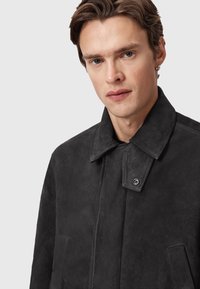 Jeune homme à la peau claire et aux cheveux bruns courts, portant une veste en daim noire avec un col et des boutons-pression, sur un fond neutre.