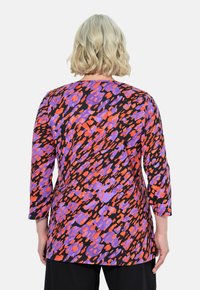 Kolmveerand varrukatega topp, mis on valmistatud pehmest kangast, millel on elav abstraktne muster oranži, lilla ja musta värviga ning ümar kaelus.