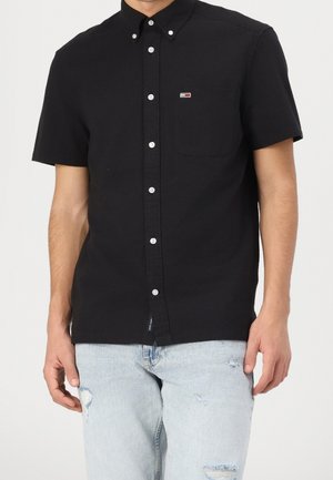 Mann trägt ein schwarzes Kurzarm-Hemd mit Knopfleiste und einem kleinen Logo auf der Brusttasche sowie hellblaue, zerrissene Jeans.