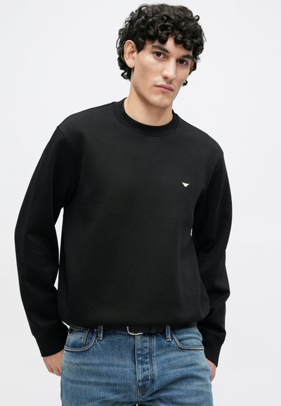 Čierna mikina s dlhými rukávmi a okrúhlym výstrihom, s malým bielym logom na hrudi. Kombinovaná s modrými džínsami.