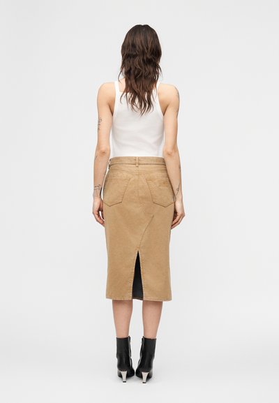 Γυναίκα με μακριά καστανά μαλλιά, που φοράει λευκό τοπ, μπεζ μίντι τζιν φούστα με σκίσιμο στο πίσω μέρος και μαύρα μποτάκια με τακούνι, γυρισμένη προς τα πίσω.