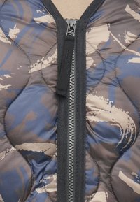 Pihe-puha mellény kék és barna terepmintával. Középen fekete cipzárral és texturált anyaggal.