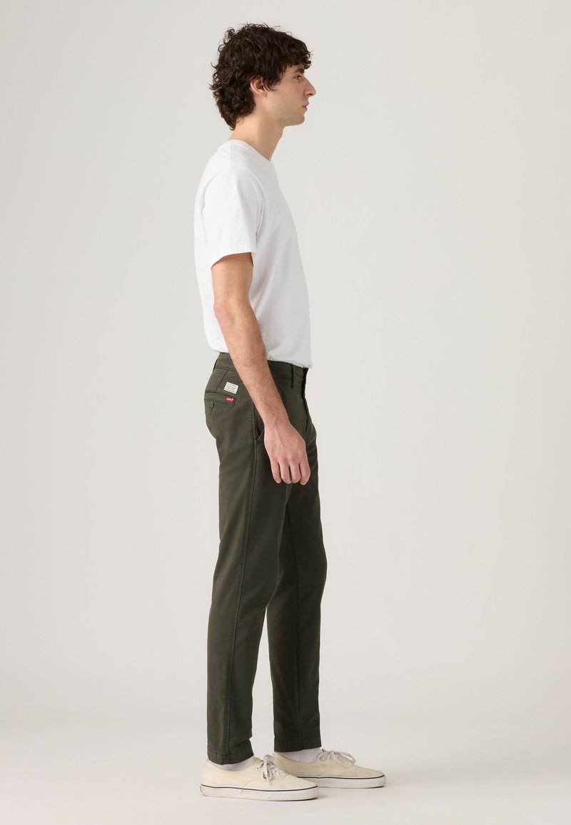 Jeune homme debout de profil portant un t-shirt blanc, un pantalon vert olive avec des étiquettes sur les poches arrière, des chaussettes blanches et des chaussures en toile écru.