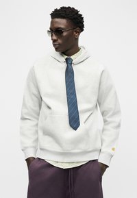 Άνδρας με γυαλιά ηλίου, ανοιχτό γκρι φούτερ με κουκούλα πάνω από πουκάμισο με μπλε ριγέ γραβάτα και μωβ παντελόνι, με τα χέρια στις τσέπες, κοιτάζει προς τα αριστερά.