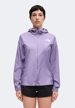 Persoon draagt een lichtpaarse capuchontrui met rits met een wit logo op de borst en zwarte shorts, staand tegen een effen achtergrond.