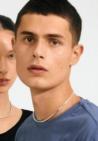 Sudraba metāla chokers kaklarota, kuru valkā modele, ar gludu, noapaļotu dizainu. Modele ir uzvilkusi zilu kreklu ar apaļu kakla izgriezumu.