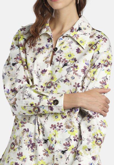 Femme portant un chemisier blanc à fleurs jaunes, violettes et brunes, manches longues, col et détails de boutons à l'avant.