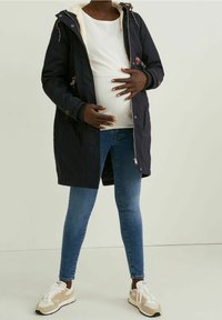 Parka azul marino con un interior de felpa y cintura ajustada, combinada con jeans ajustados azules y zapatillas beige. Cuenta con cierre de cremallera y botón.