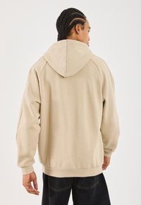 Person med fletninger iført en løs beige hættebluse og sorte bukser, vendt væk fra en ensfarvet hvid baggrund.