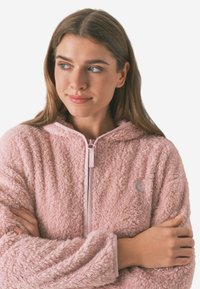 Lyserød, fluffy hoodie med tekstureret fleece materiale, lynlås og hætte. Accentueret med en lys rosa kant og et lille logo.