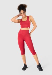 Soutien-gorge de sport rouge et leggings capri taille haute en tissu stretch. Les accessoires comprennent des chaussures de sport noires et des détails de marque minimalistes.