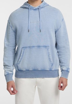 Hellblauer Guess Jeans Hoodie mit Fronttasche und Kordeln, getragen zu weißen Hosen, dargestellt vom Hals bis zu den Hüften.
