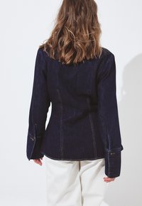 Persoon met golvend bruin haar, gekleed in een donkerblauwe spijkerjack en witte broek, staande tegen een effen lichte achtergrond, met de rug naar de camera gekeerd.