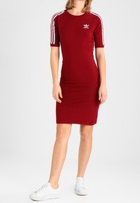 Czerwona dopasowana sukienka z krótkimi rękawami, białe paski Adidas na ramionach oraz małe logo na piersi. Gładki materiał jersey, do kolan.