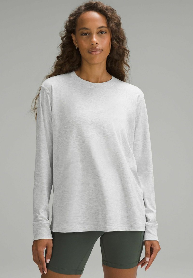 Jasnoszara, długosleeve'owa koszula z miękkiego, teksturowanego materiału, z okrągłym dekoltem i luźnym krojem, zestawiona z ciemnozielonymi szortami.