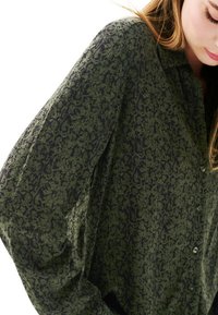 Chemise verte à motif avec une coupe ample, des manches longues et une fermeture à boutons. Le tissu a une texture douce avec un motif floral foncé.