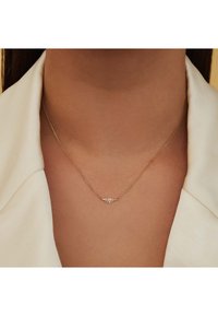 Gouden ketting met een verfijnde bladvormige hanger, met verfijnde details, hangend aan een dunne ketting. Draagt tegen een lichtgekleurde kraag.