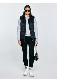 Naine kannab musti päikeseprille, musta vattvesti, halli kapuutsiga pusa, musti pükse, valgeid tossusid triibuliste sokkidega ning hoiab käes musta käekotti kuldse ketiga.