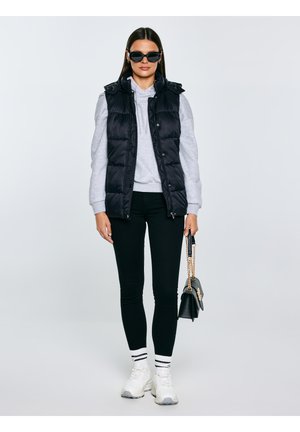 Kvinde iført sorte solbriller, sort dunvest, grå hættetrøje, sorte bukser, hvide sneakers med stribede sokker, holder en sort håndtaske med guldkæde.