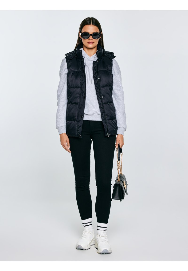 Naine kannab musti päikeseprille, musta vattvesti, halli kapuutsiga pusa, musti pükse, valgeid tossusid triibuliste sokkidega ning hoiab käes musta käekotti kuldse ketiga.