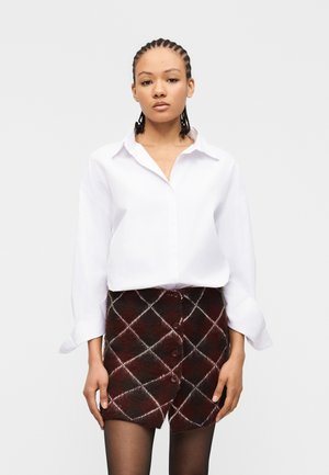 Jeune femme aux cheveux tressés portant une chemise blanche boutonnée, une jupe à carreaux bordeaux et noire boutonnée, et des collants noirs, debout devant un fond blanc.