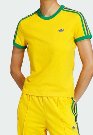 Osoba ubrana w żółty strój sportowy Adidas z zielonymi i niebieskimi paskami na rękawach i spodniach, ręce spoczywają za plecami.