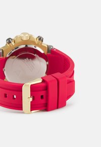 Rotes Silikon-Uhrenarmband mit goldenem Schnallen- und Hardware-Elementen, ausgestattet mit einer glatten Oberfläche und einem runden Gehäuse aus Edelstahl.