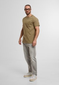 T-shirt khaki a maniche corte con tasca sul petto, abbinato a pantaloni dritti grigio chiaro e sneakers beige. Tessuto texturizzato, vestibilità rilassata.