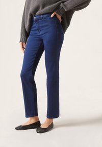 Siniset suoralahkeiset farkut keskikorkealla vyötäröllä, joissa on edessä nappikiinnitys ja perinteiset taskut. Päällä mustat tasapohjaiset kengät.