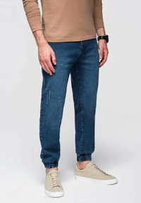 Camisa de manga larga marrón claro, pantalones jogger de mezclilla azul con puños elásticos, zapatillas de deporte beige claro, mostrando un atuendo informal.