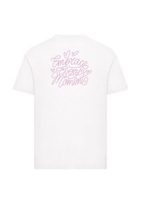 Camiseta blanca con texto en cursiva rosa en la parte posterior que dice "Abrazar Cada Momento", con acentos de corazones alrededor del texto.
