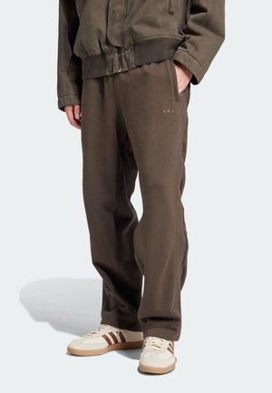 Person trägt braune Adidas-Sweathose und passende Jacke, weiße Sneaker mit braunen Streifen, steht mit einer Hand in der Tasche.