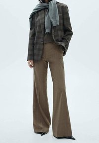 Geruite blazer in donkere tinten, gecombineerd met bruin gestreepte broeken. Zachte grijze sjaal over een aangesloten trui gedrapeerd. Glanzende zwarte hakken.