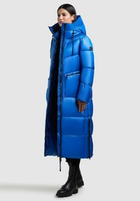 Langer Puffer-Mantel in leuchtendem Blau, mit hohem Kragen, Reißverschlusstaschen und glänzender Textur. Kombiniert mit schwarzen Knöchelstiefeln.