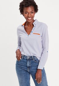 Chemise boutonnée blanche avec des motifs géométriques bleus, col orange contrastant et détails de poche. Assortie à un jean bleu taille haute.